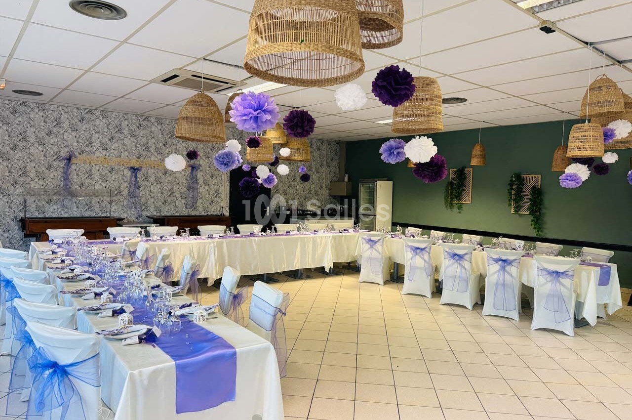Salle décorée pour un événement avec tables dressées, nappes blanches, chemins de table violets et suspensions en osier.