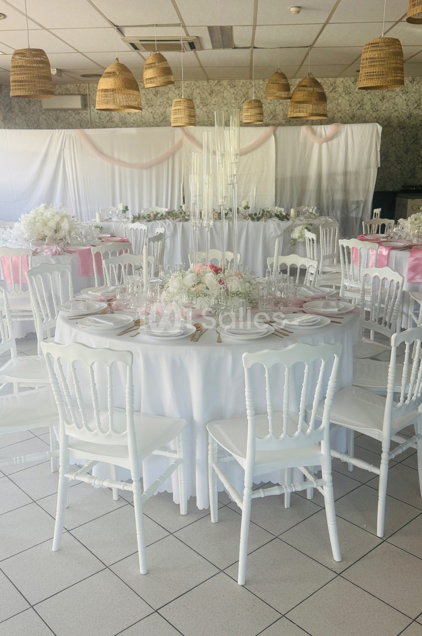 Salle de réception décorée avec des tables rondes dressées, nappes blanches, chaises blanches et touches de rose.