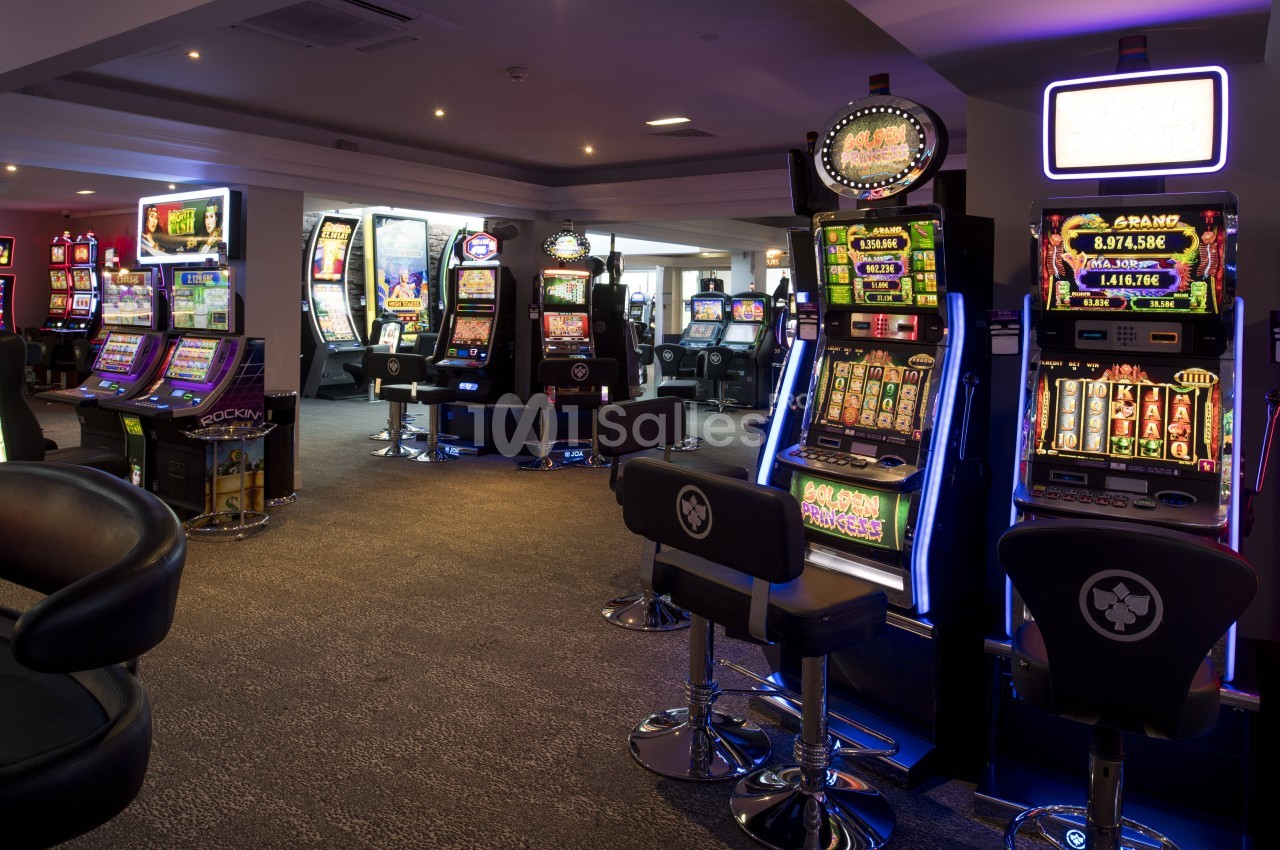 Salle de jeux avec plusieurs machines à sous illuminées et des sièges devant chaque appareil.