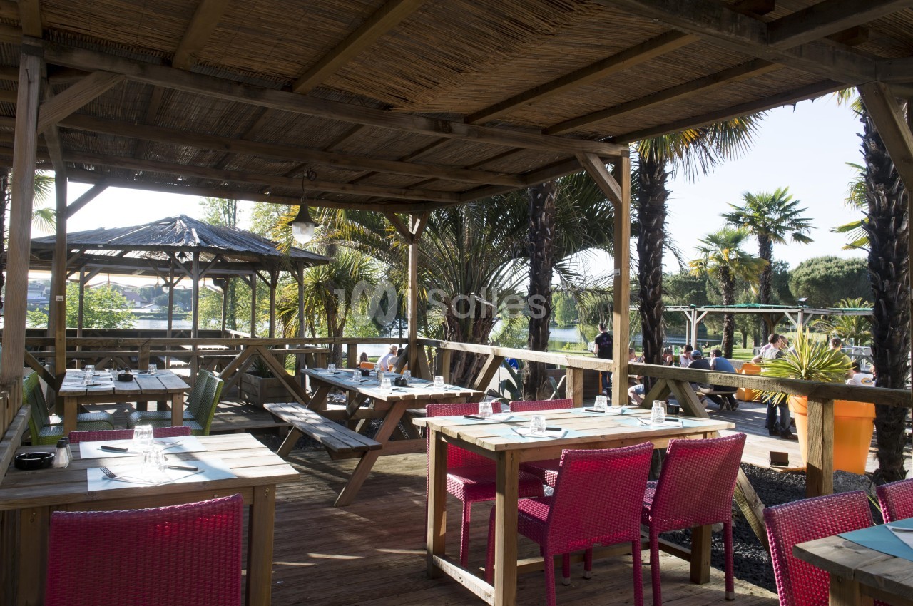 Terrasse en bois ombragée avec tables dressées, chaises colorées et vue sur un jardin avec palmiers.