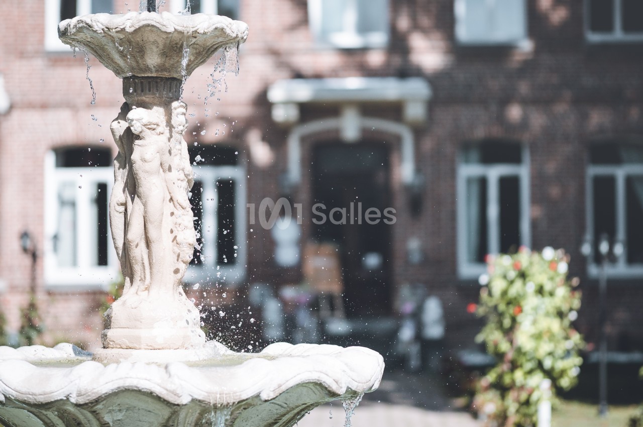 Fontaine en pierre ornée de sculptures, avec en arrière-plan une façade en briques et un jardin fleuri.