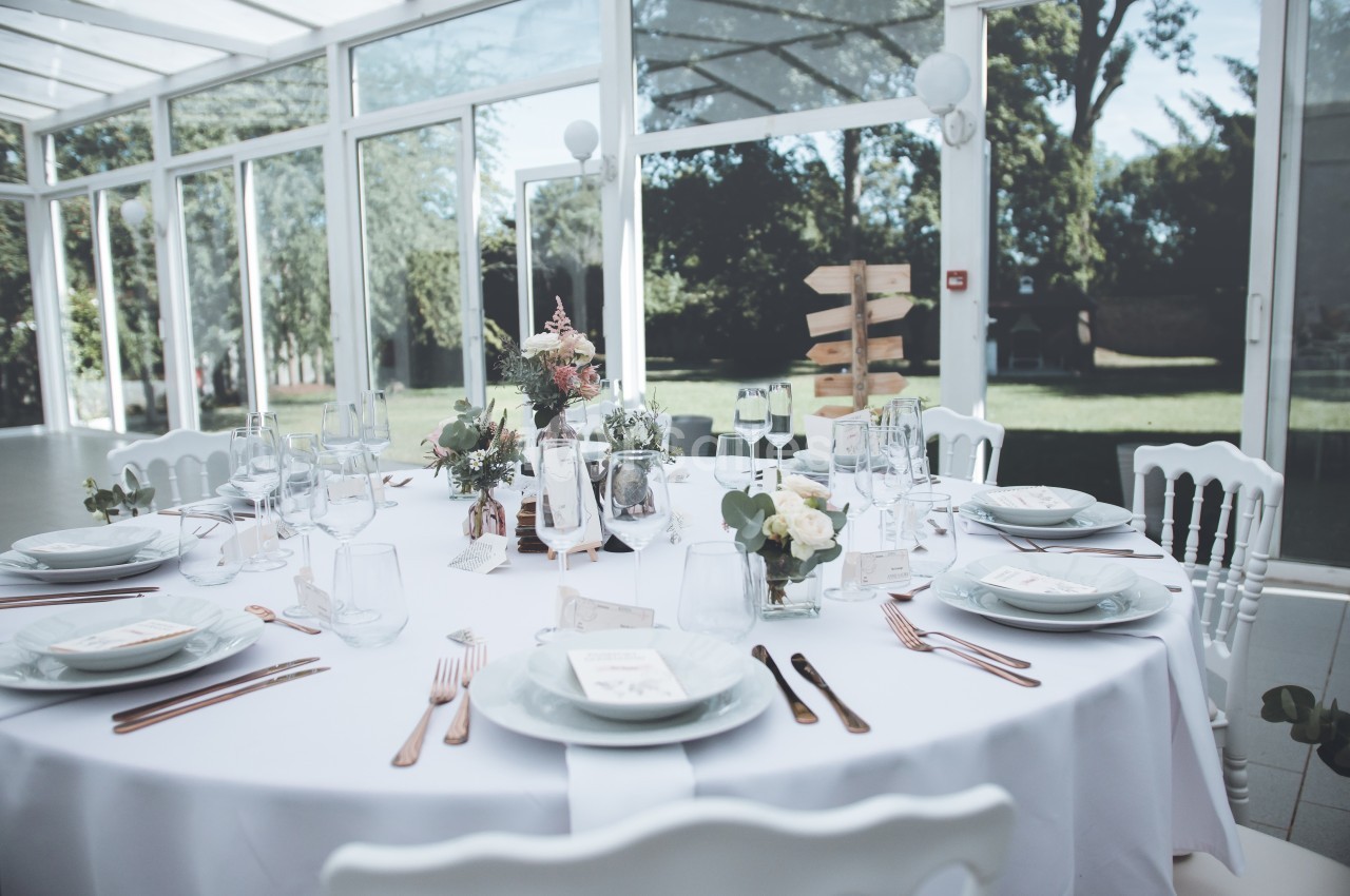 Table ronde élégamment dressée avec vaisselle blanche, décorations florales et vue sur un jardin à travers de grandes baies…