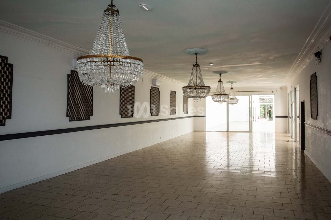 Salle vide avec carrelage clair, murs blancs, lustres en cristal suspendus et lumière naturelle entrant par une porte vitrée.