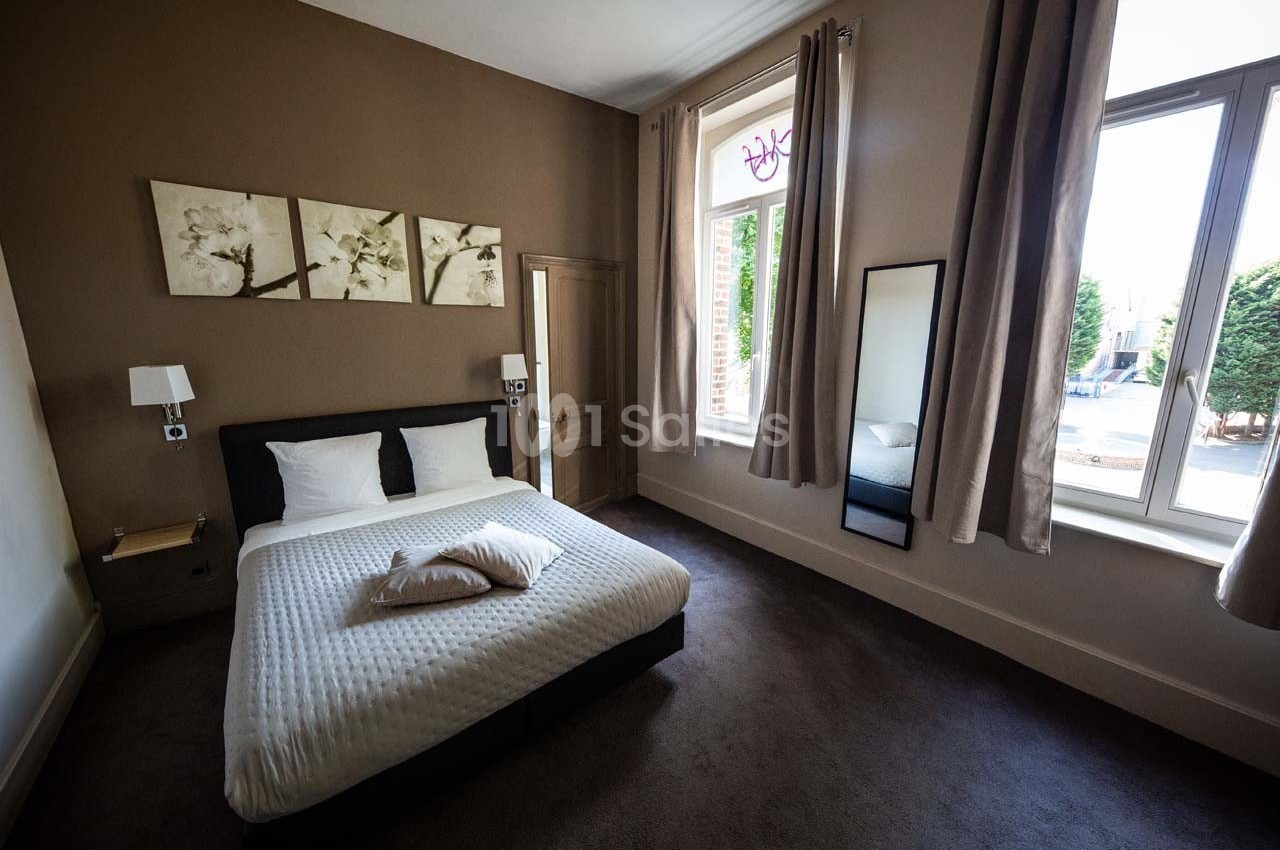 Chambre avec lit double, murs beige, grande fenêtre avec rideaux, miroir en pied et décorations murales florales.