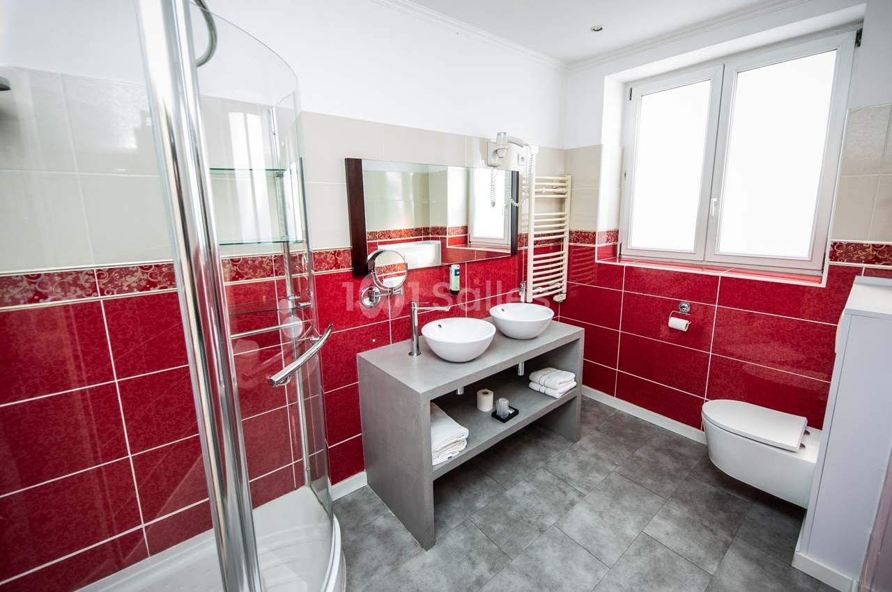 Salle de bain moderne avec douche vitrée, double vasque, carrelage rouge et blanc, et fenêtre apportant de la lumière…