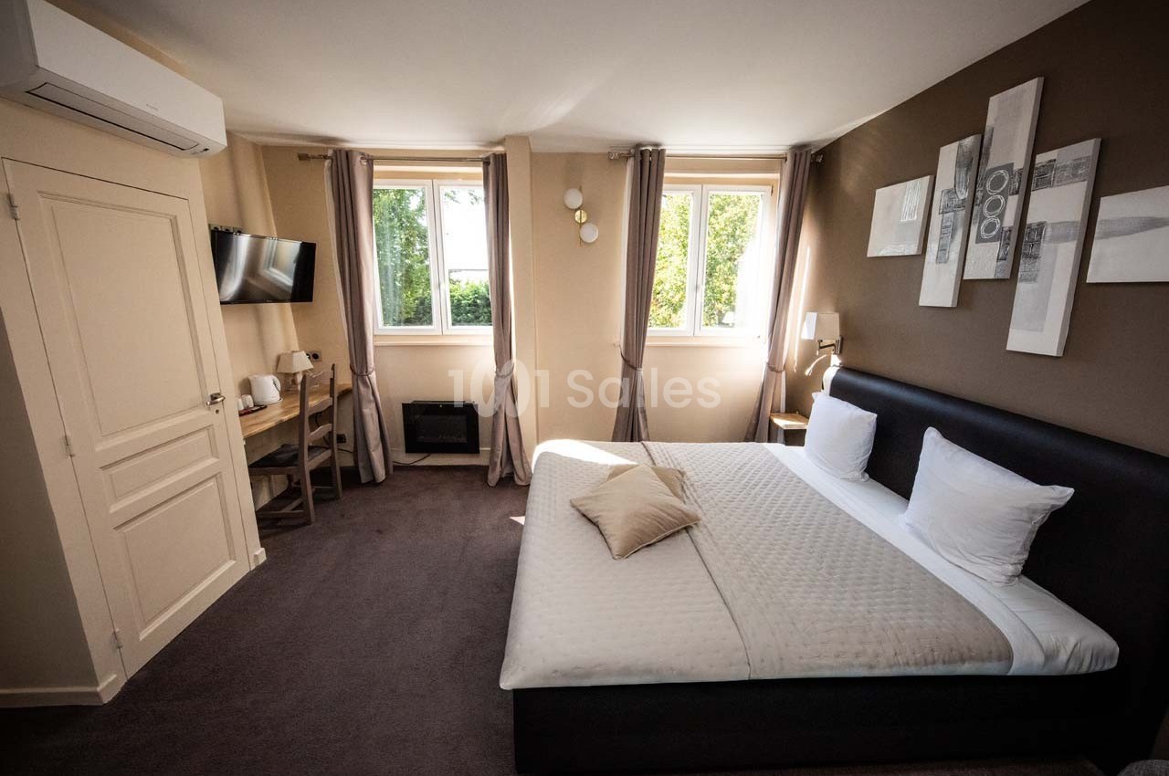 Chambre lumineuse avec lit double, bureau, télévision murale, fenêtres donnant sur un espace vert et décoration moderne.
