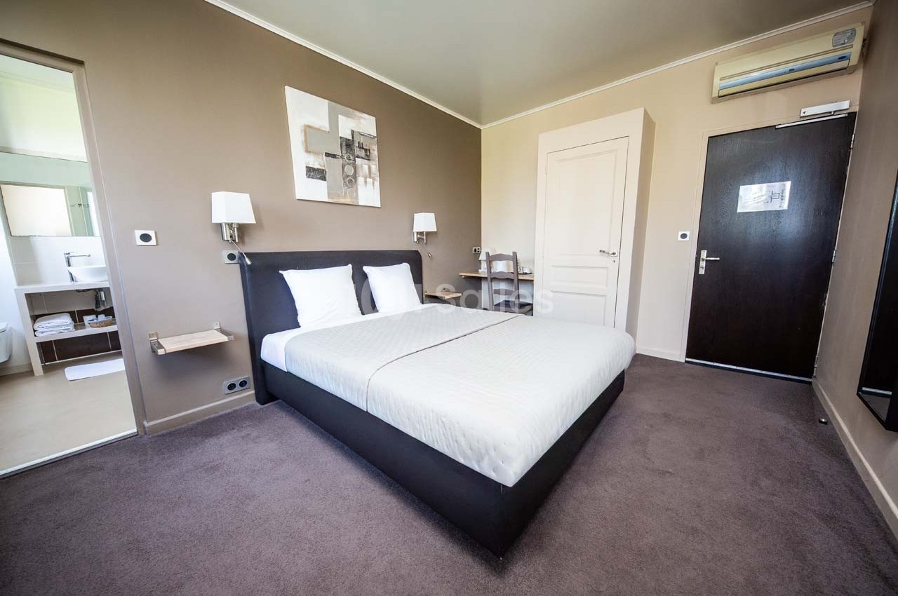Chambre d'hôtel avec lit double, murs beige, bureau, chaise, porte noire et salle de bain visible en arrière-plan.