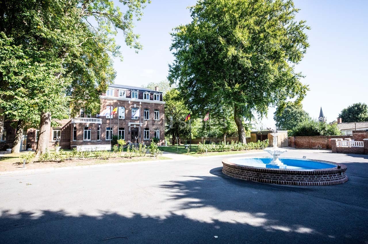 Bâtiment en briques entouré d'arbres avec une fontaine circulaire au premier plan, sous un ciel dégagé.