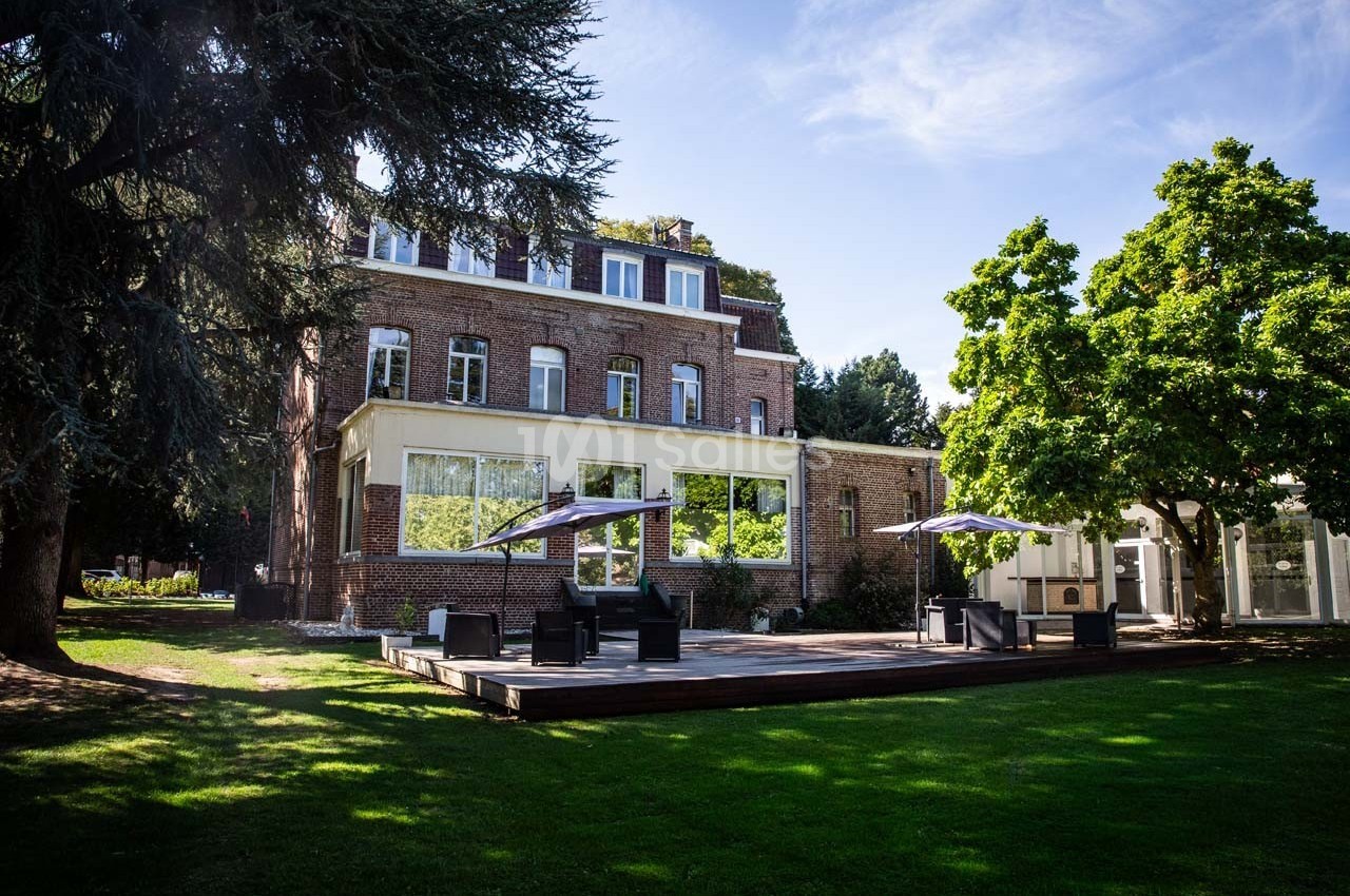 Façade d'un bâtiment en briques entouré d'arbres, avec une terrasse en bois aménagée de tables et parasols.