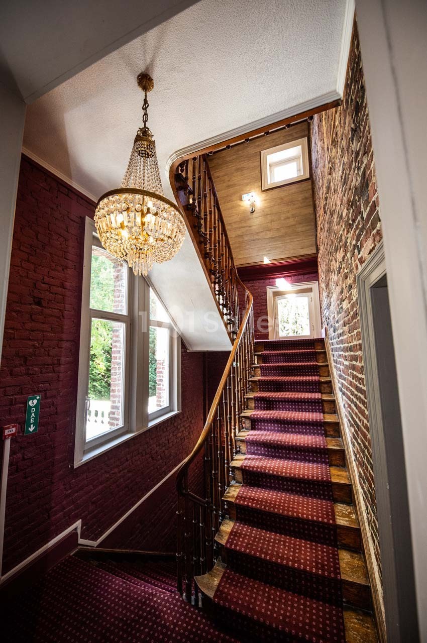 Escalier en bois avec tapis rouge, murs en briques apparentes, lustre en cristal et lumière naturelle provenant des fenêtres.