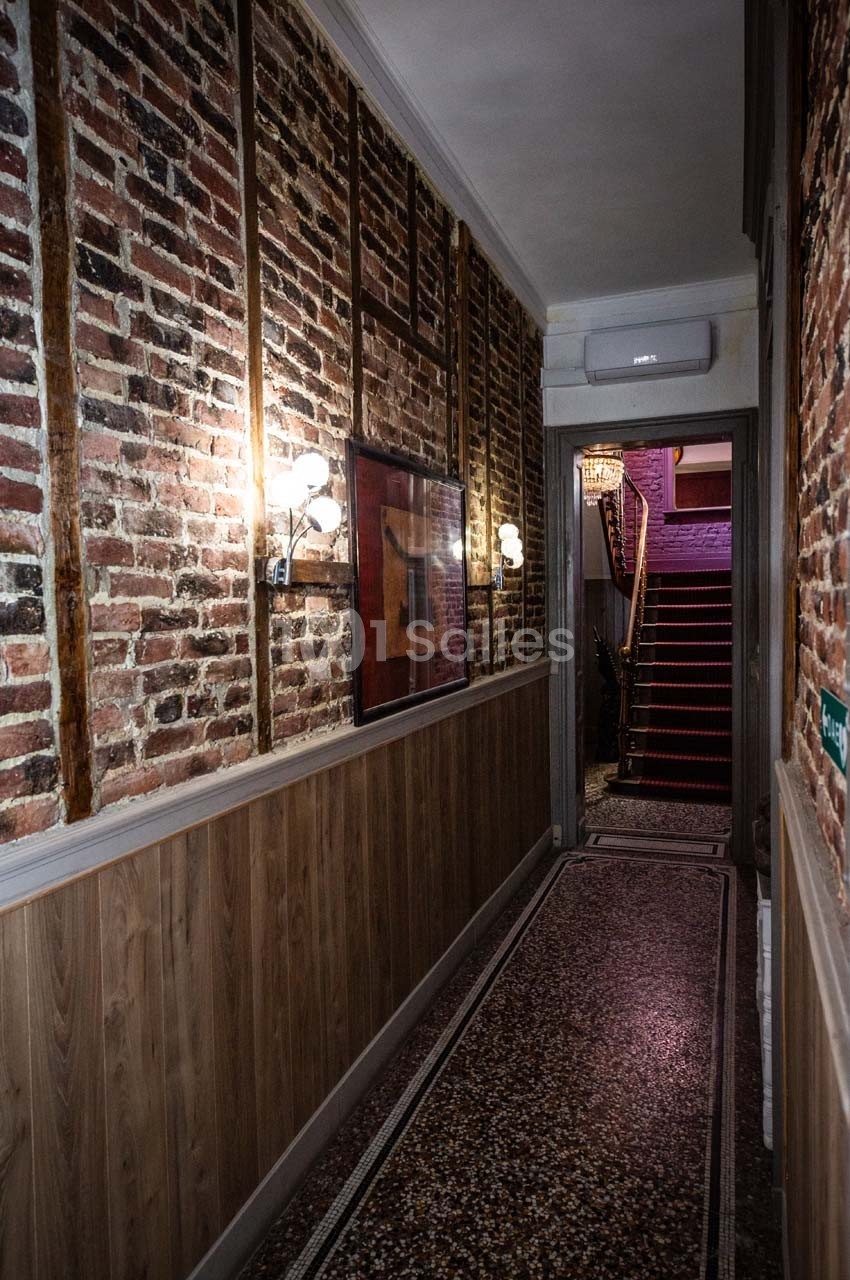 Couloir étroit avec murs en briques et bois, sol en terrazzo, éclairage mural et escalier au fond.