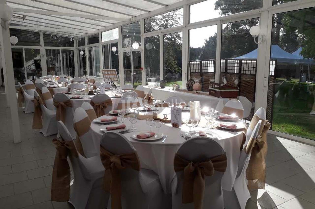 Salle lumineuse avec tables rondes décorées pour un événement, vue sur un jardin à travers de grandes baies vitrées.