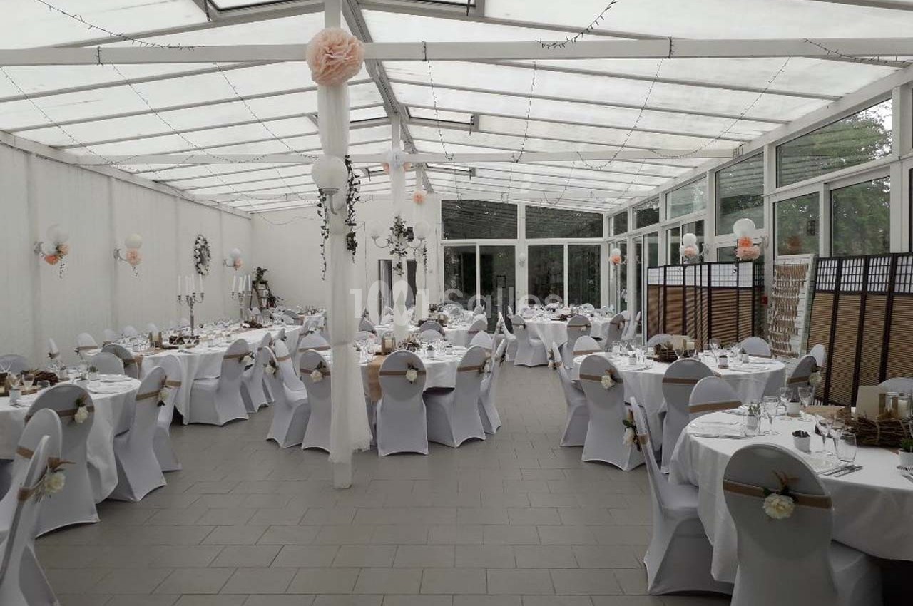 Salle de réception décorée avec des tables rondes dressées, chaises blanches ornées de nœuds et éclairage naturel.