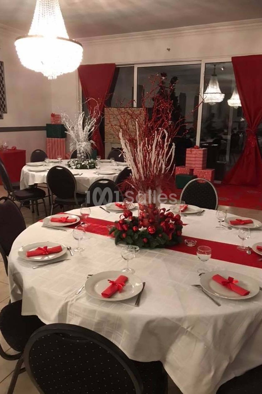 Salle décorée pour un repas festif, avec tables dressées, nappes blanches, décorations rouges et chandeliers.
