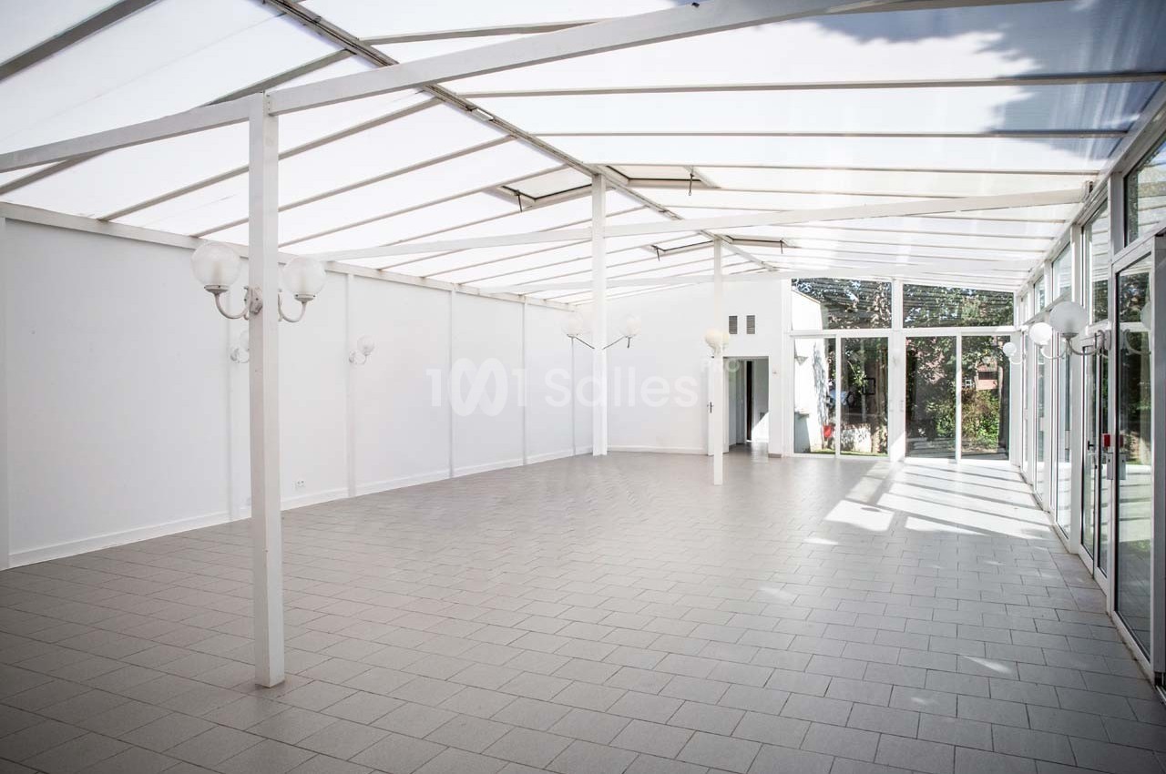 Salle lumineuse avec toit en verre, murs blancs, sol carrelé gris et grandes baies vitrées donnant sur un jardin.