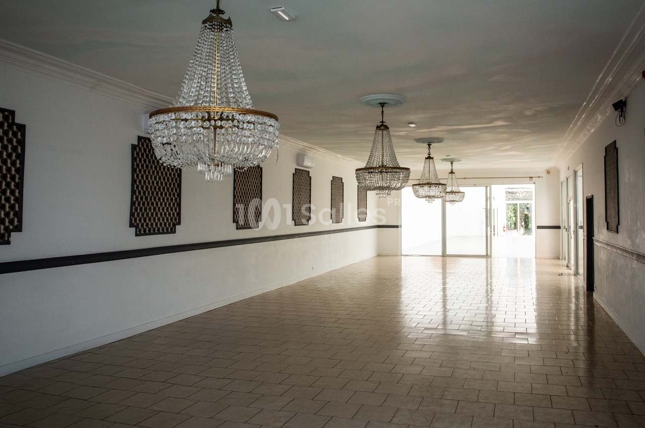 Salle vide avec carrelage clair, murs blancs, lustres en cristal suspendus et lumière naturelle entrant par une porte vitrée.