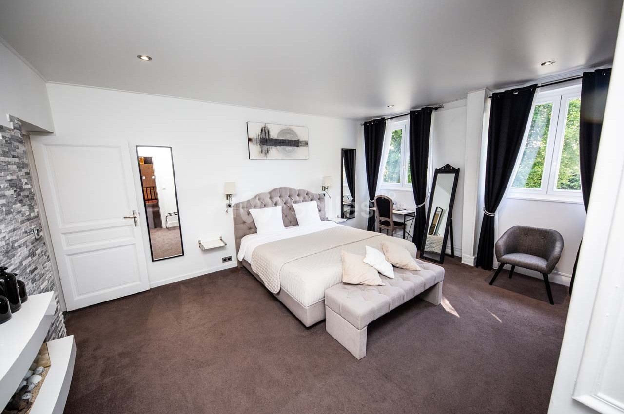 Chambre lumineuse avec lit double, fauteuil, miroirs, rideaux noirs et fenêtres donnant sur un espace vert.