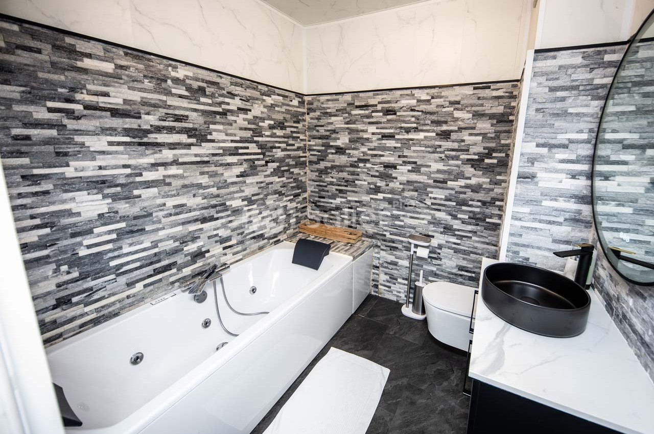 Salle de bain moderne avec baignoire à jets, lavabo noir, bidet et murs en carrelage gris texturé.