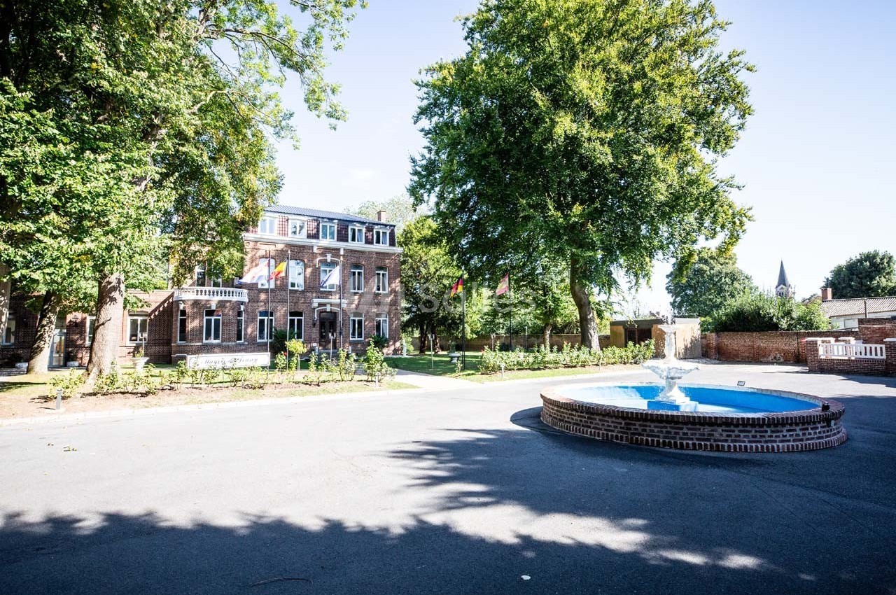 Bâtiment en briques entouré d'arbres avec une fontaine circulaire au premier plan, sous un ciel dégagé.