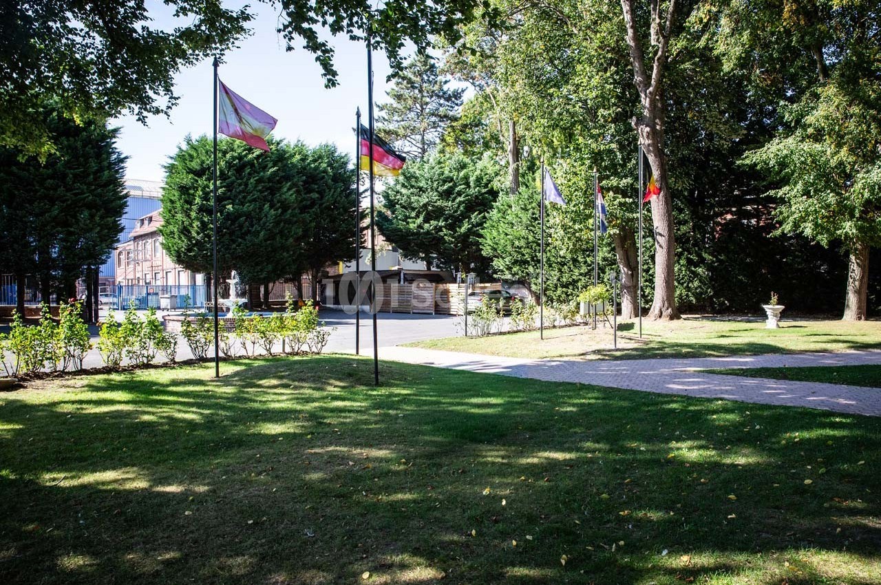 Pelouse avec arbres, allée pavée et drapeaux sur mâts devant des bâtiments en arrière-plan.