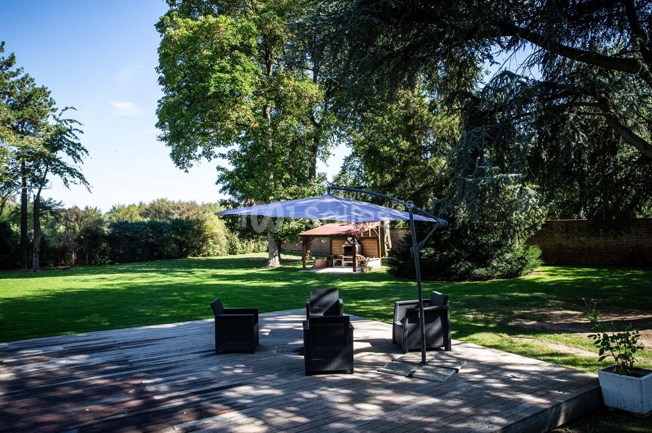 Terrasse en bois avec fauteuils noirs sous un parasol, entourée d'un grand jardin arboré par une journée ensoleillée.
