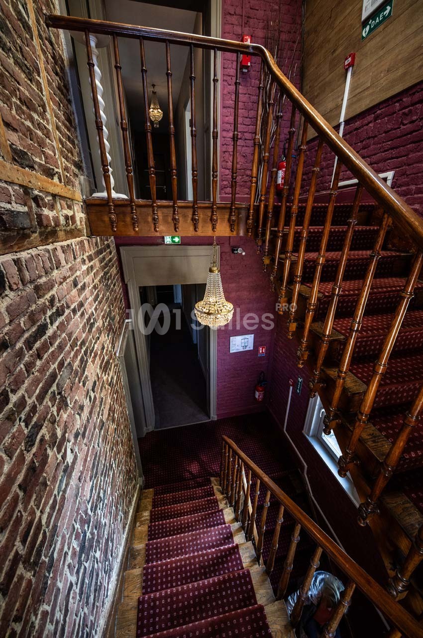 Escalier en bois avec tapis rouge, murs en briques apparentes et lustre suspendu dans un bâtiment ancien.