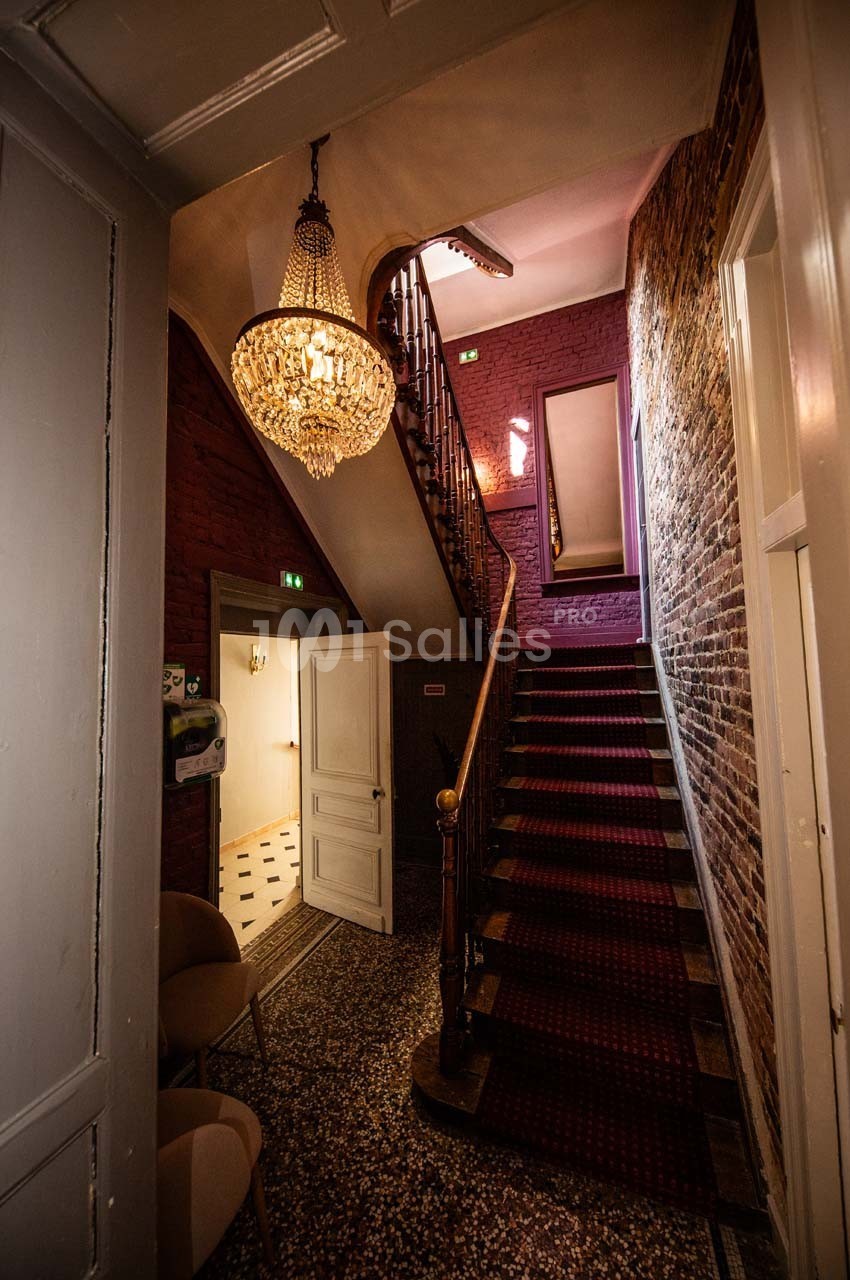 Escalier en bois avec tapis rouge dans un intérieur aux murs en briques, éclairé par un lustre imposant.