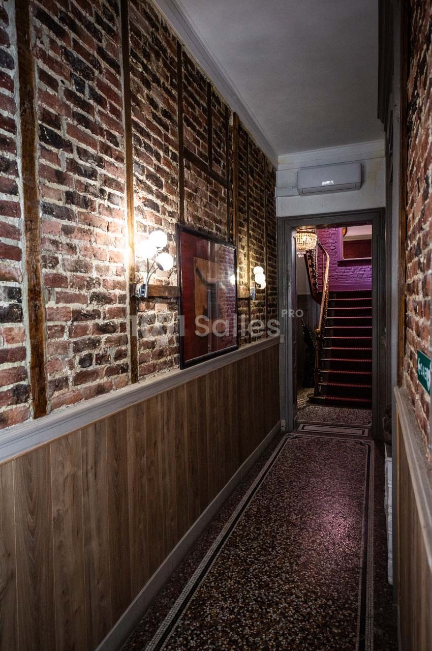 Couloir étroit avec murs en briques et bois, sol en terrazzo, éclairage mural et escalier au fond.
