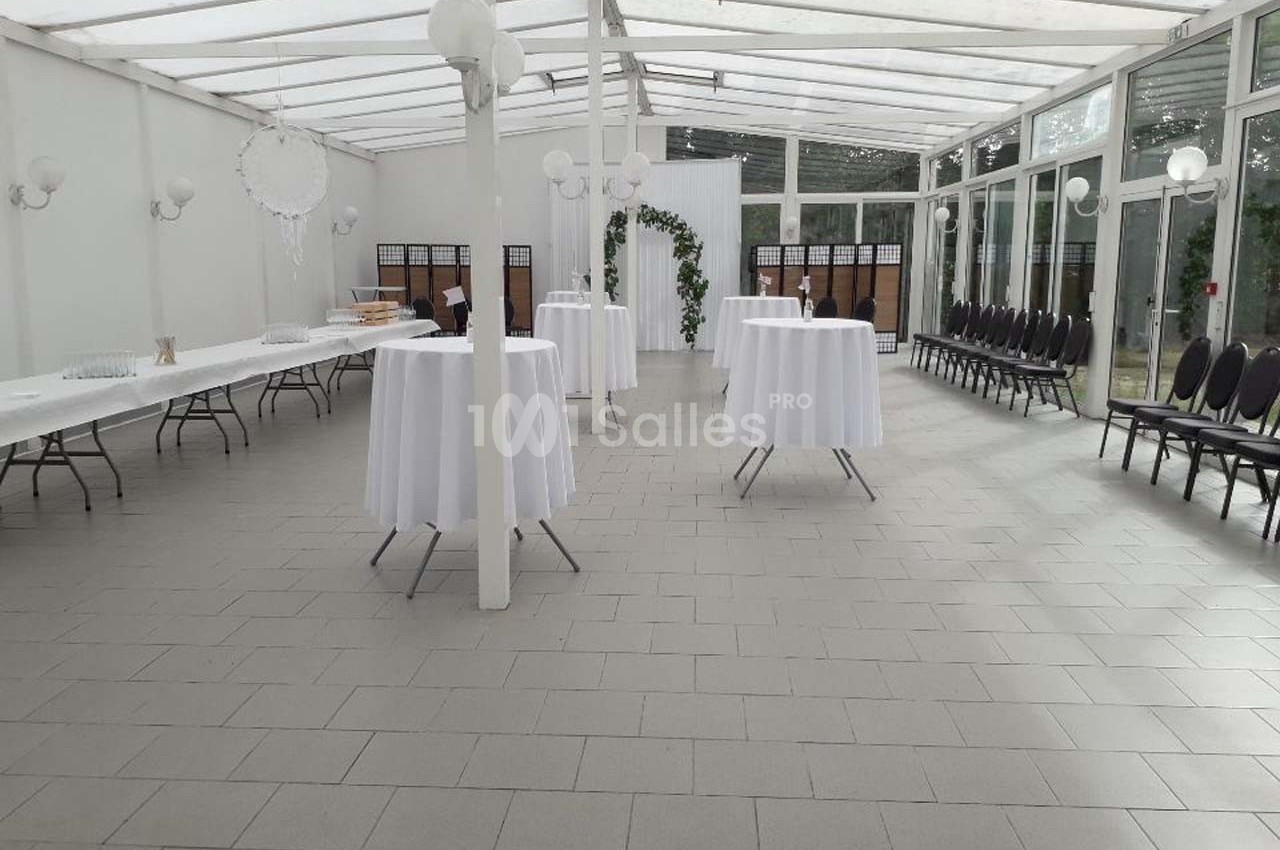 Salle lumineuse avec tables hautes drapées de blanc, chaises alignées et décoration végétale au fond.