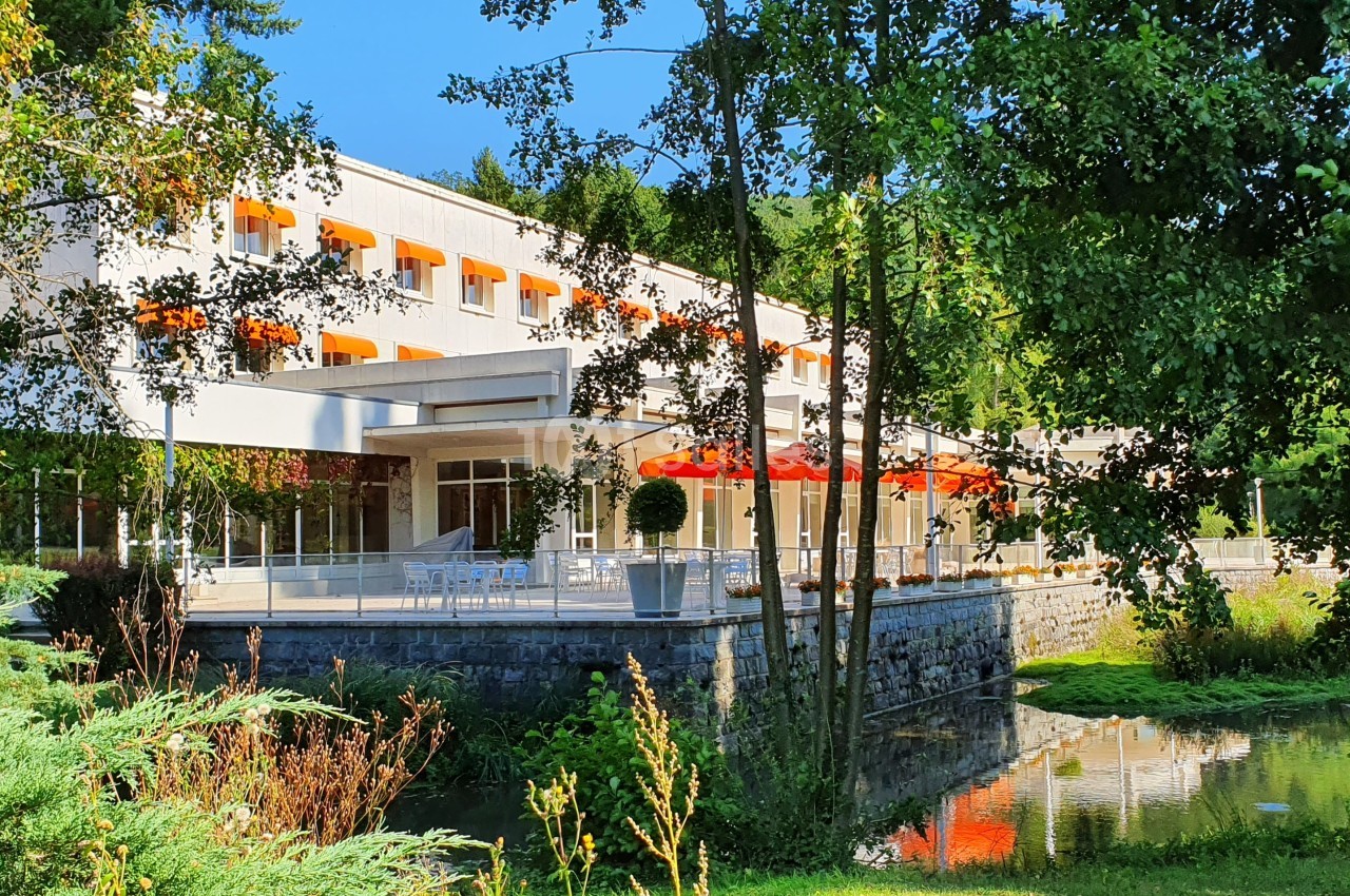 Bâtiment moderne entouré de verdure, avec des baies vitrées, des parasols rouges et un petit plan d'eau au premier plan.