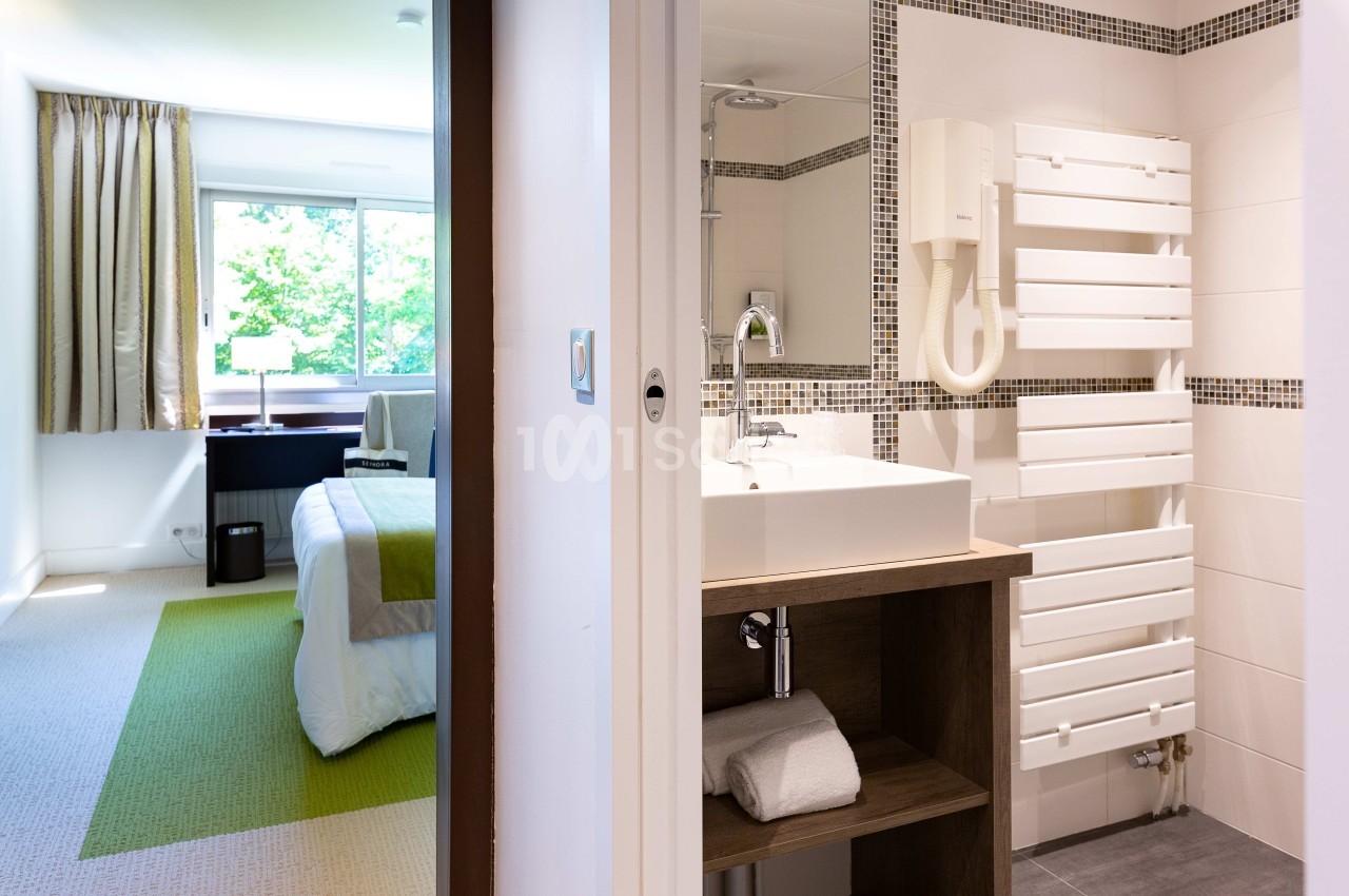 Vue d'une chambre d'hôtel lumineuse avec salle de bain attenante, lavabo, sèche-serviettes et lit près d'une fenêtre.