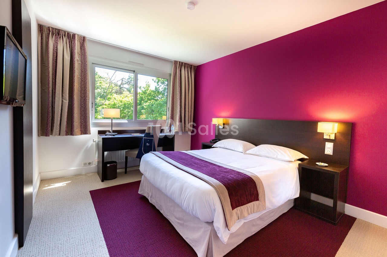 Chambre d'hôtel lumineuse avec un lit double, un bureau près d'une fenêtre et des murs partiellement violets.