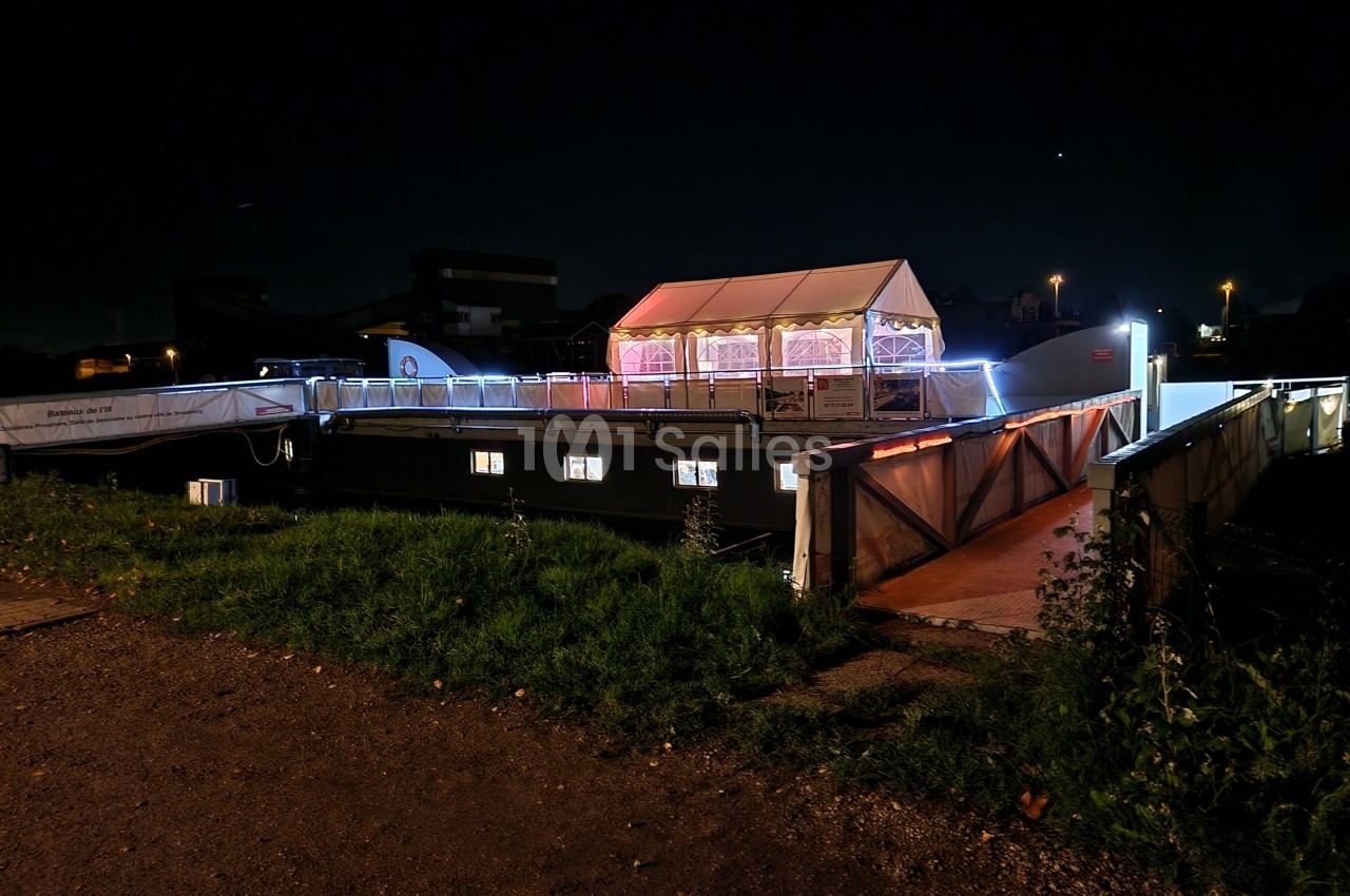 Bateau illuminé amarré de nuit, avec une tente blanche éclairée sur le pont et un chemin d'accès en premier plan.