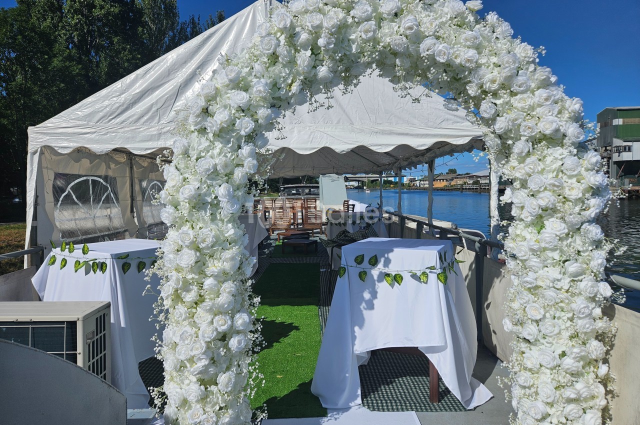 Arche de fleurs blanches devant une tente sur un bateau, avec vue sur l'eau et des bâtiments en arrière-plan.