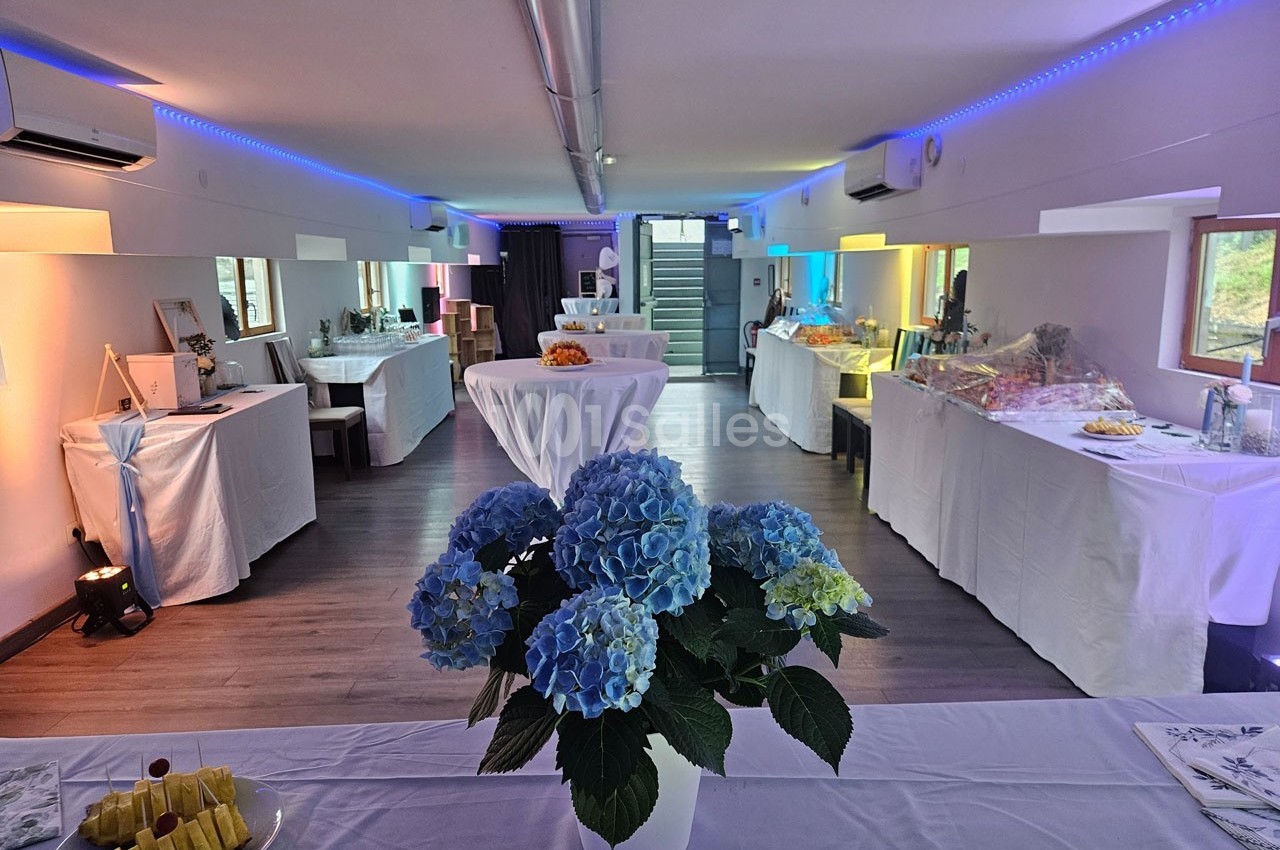 Salle décorée pour un événement, avec des tables dressées, des fleurs bleues au premier plan et un éclairage coloré.