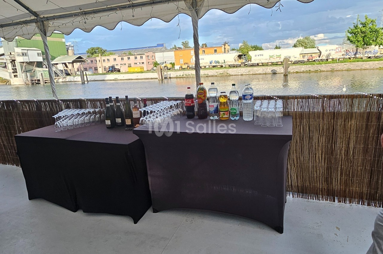 Deux tables noires avec boissons, verres et bouteilles, installées sous une tente près d'une rivière.