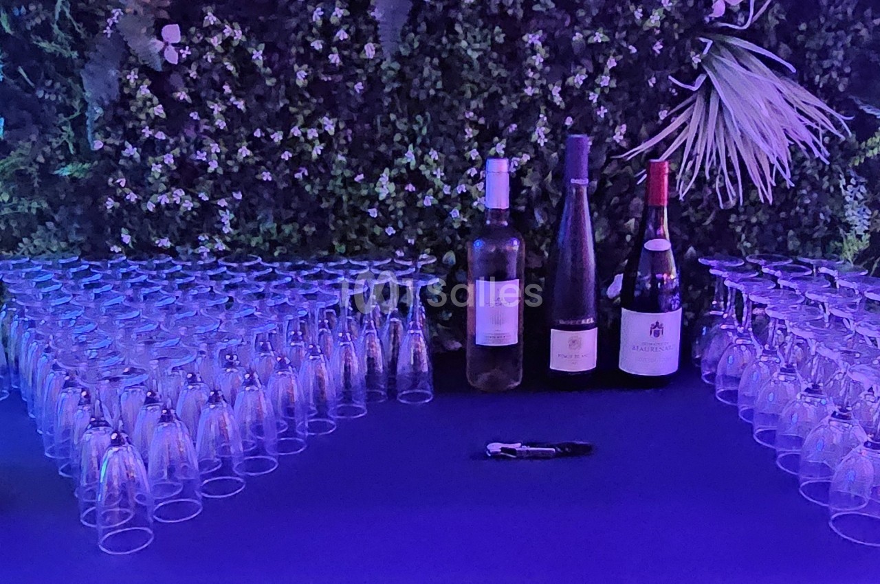 Bouteilles de vin et flûtes en verre disposées sur une table devant un mur végétal éclairé par une lumière violette.