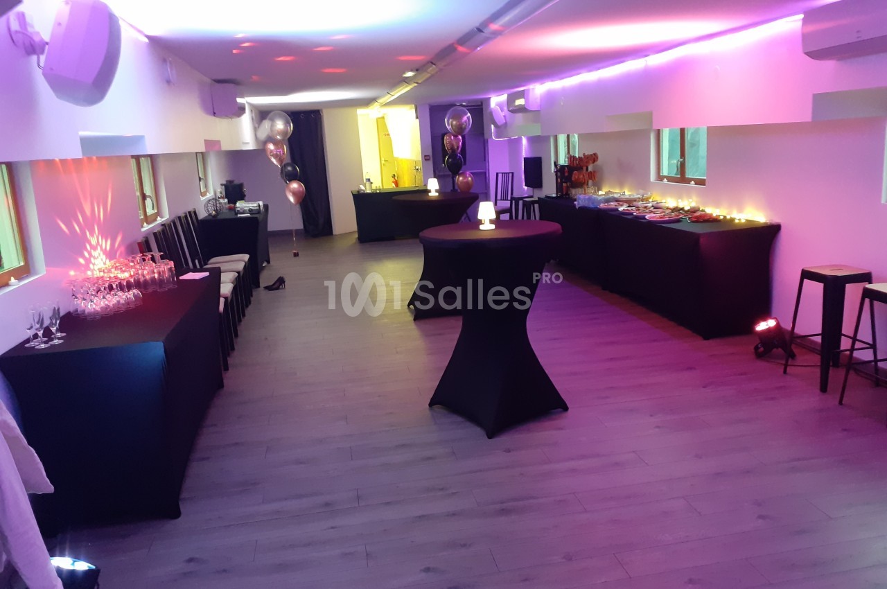 Salle décorée pour un événement avec tables hautes, buffet, ballons et éclairage coloré.