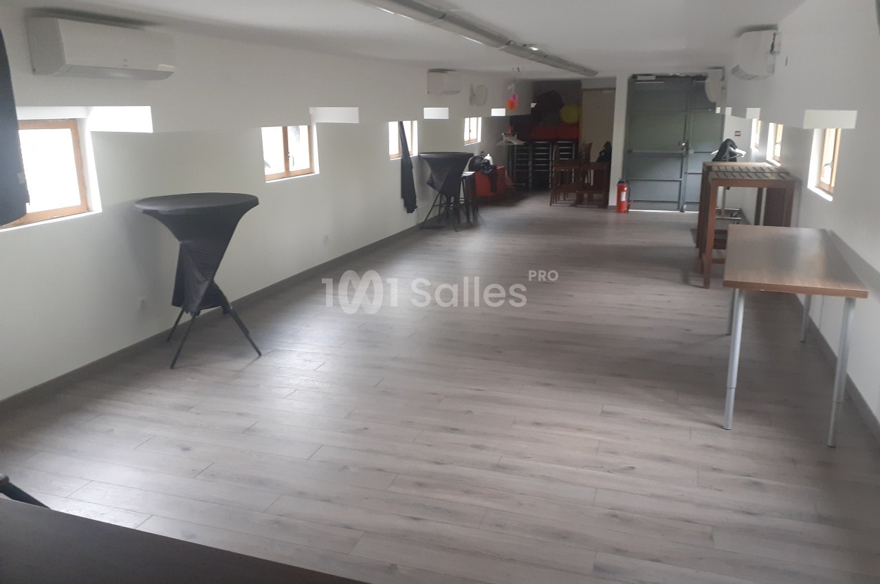 Salle lumineuse avec parquet clair, tables pliantes, chaises empilées et fenêtres laissant entrer la lumière naturelle.