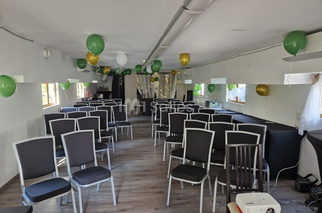Salle aménagée avec des rangées de chaises noires, décorée de ballons verts, blancs et dorés, et équipée d'un projecteur.