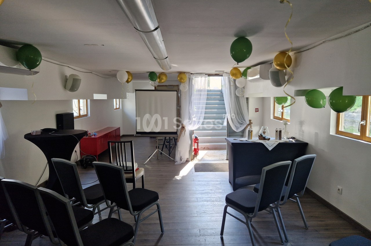 Salle décorée avec des ballons, équipée de chaises, d'un écran de projection et d'une table pour un événement.