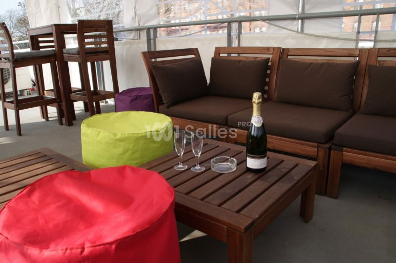 Salon extérieur avec mobilier en bois, poufs colorés, bouteille de champagne, verres et cendrier sur une table basse.