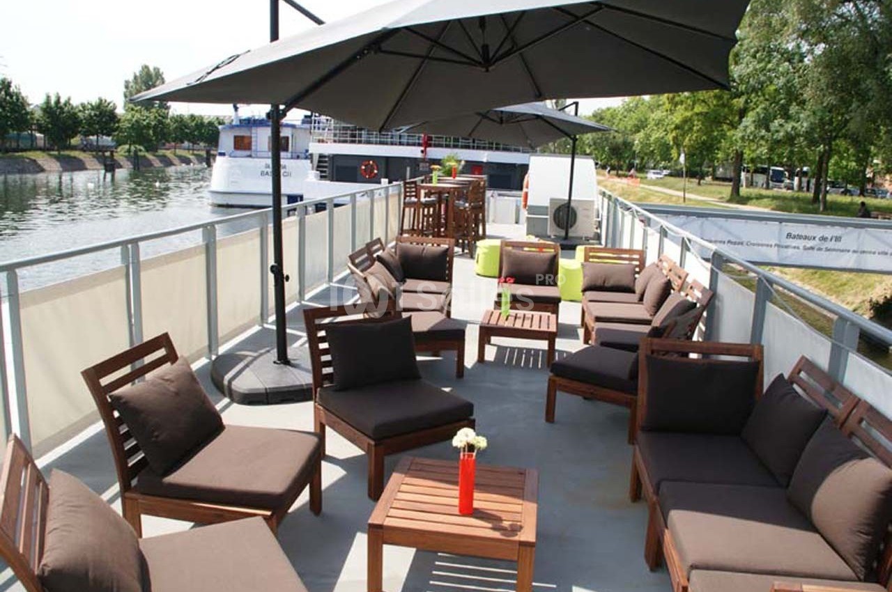 Terrasse aménagée sur un bateau avec des fauteuils en bois, des coussins marron et des parasols, au bord d'un canal.