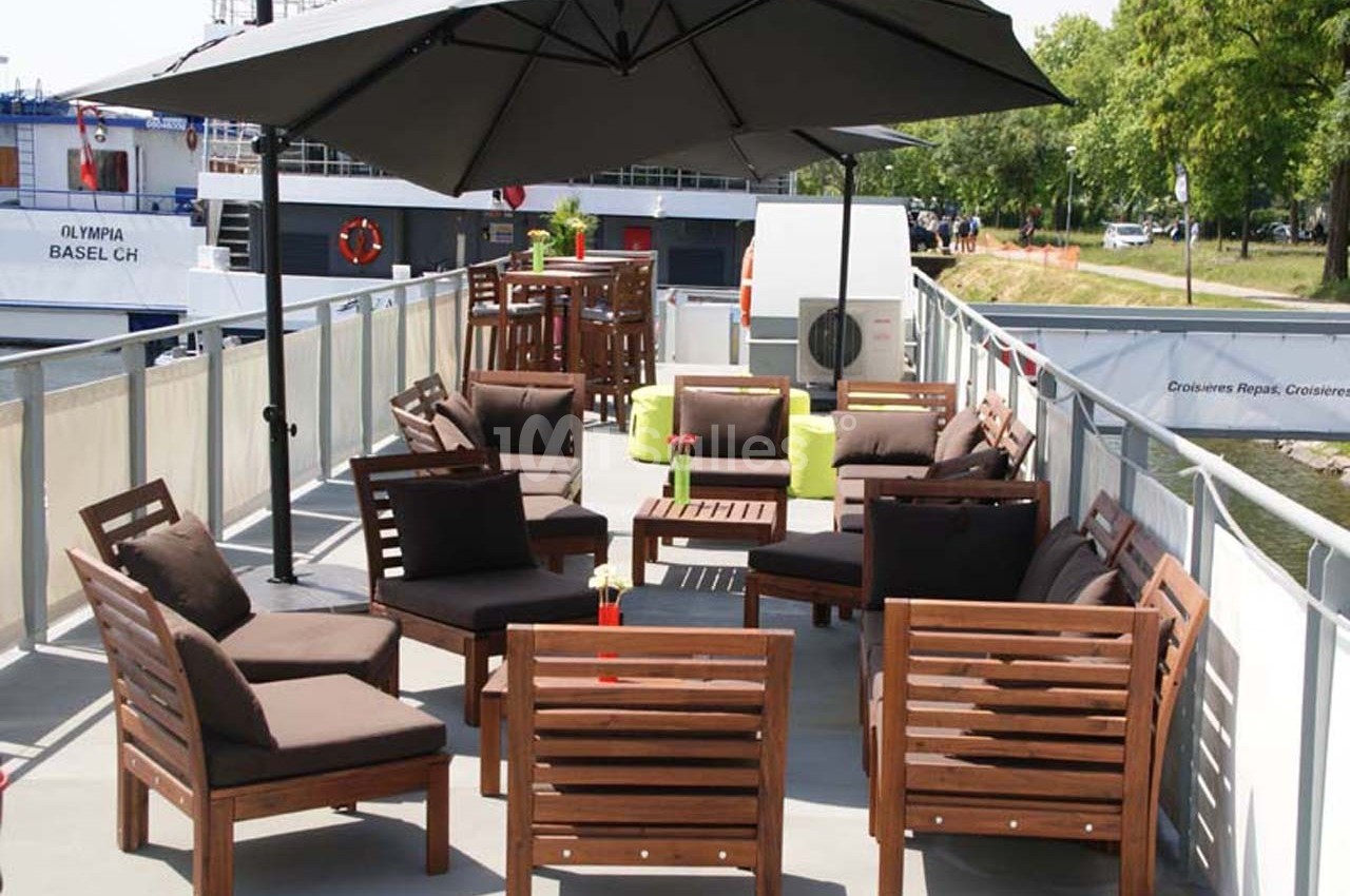 Mobilier de jardin en bois avec coussins bruns, disposé sous un grand parasol sur le pont d'un bateau.