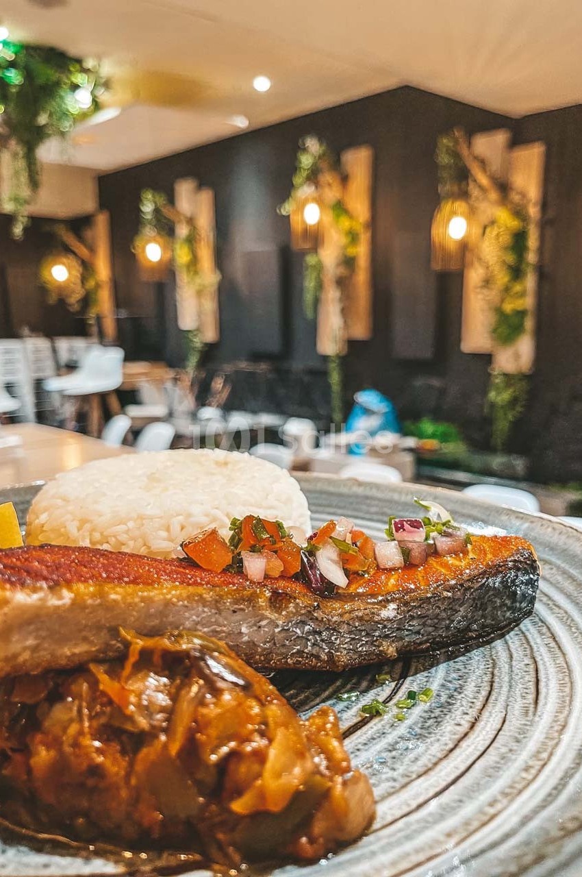 Assiette avec filet de poisson grillé, riz blanc, légumes en sauce et garniture, dans un restaurant à la décoration végétale.