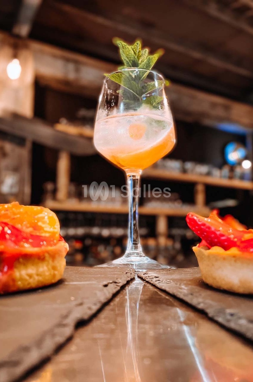 Un cocktail décoré de menthe posé sur une table, entouré de petites tartes aux fruits dans un cadre de bar chaleureux.