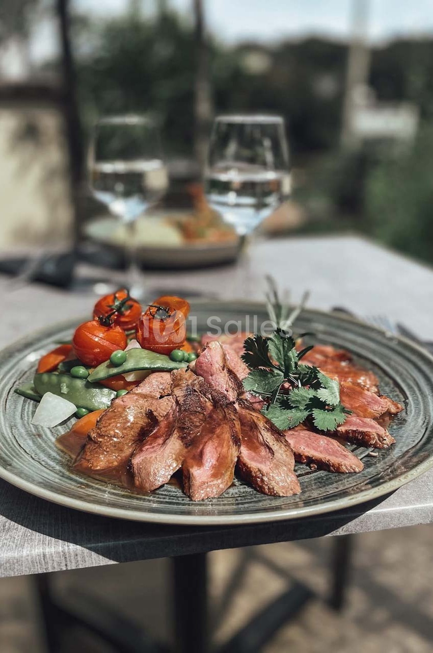 Assiette de viande grillée accompagnée de légumes colorés, servie sur une table extérieure avec des verres d'eau en arrière…