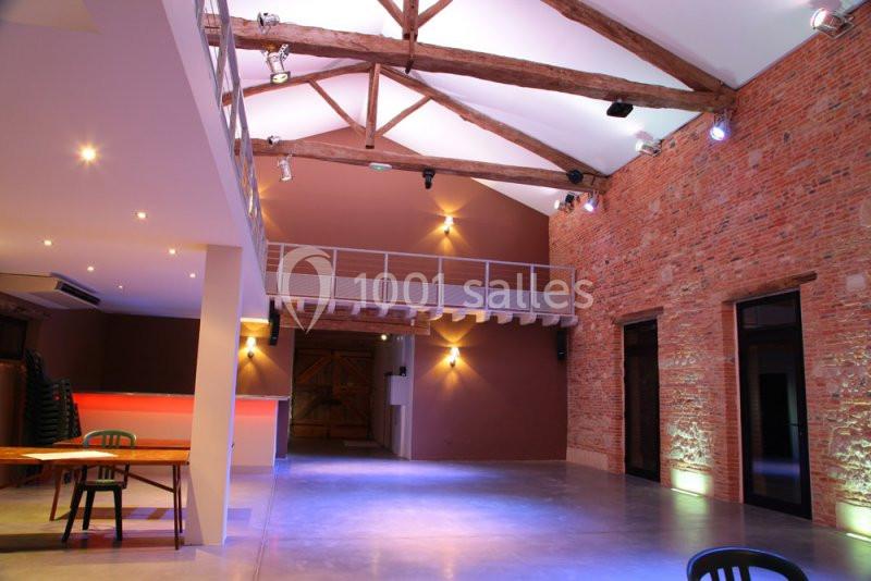 Salle spacieuse avec murs en briques, poutres apparentes, éclairage moderne et mezzanine en bois.