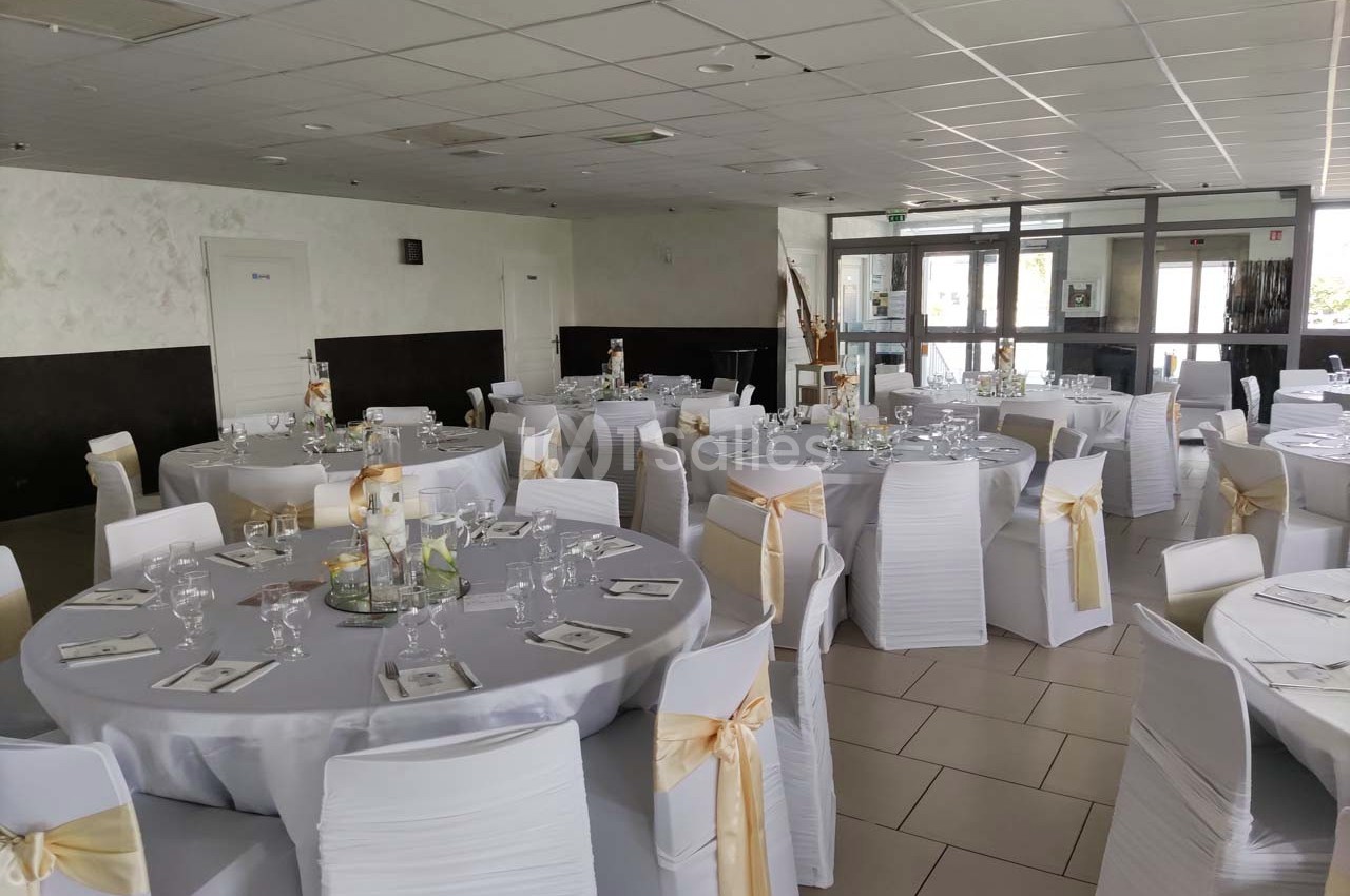 Salle de réception décorée avec des tables rondes, nappes blanches et chaises ornées de nœuds dorés.
