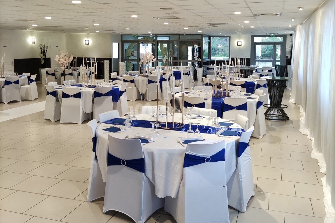 Salle de réception lumineuse avec tables rondes dressées, nappes blanches et décorations bleues.